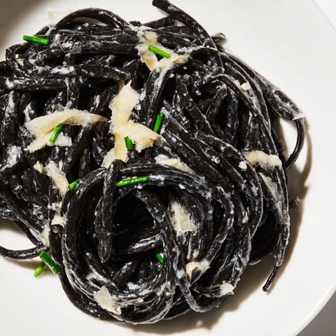 Close-up of Squid Ink Pasta Dramatic twirled on a fork, coated in silky cream sauce with visible lemon zest and fresh chives, perfect for a dinner party.