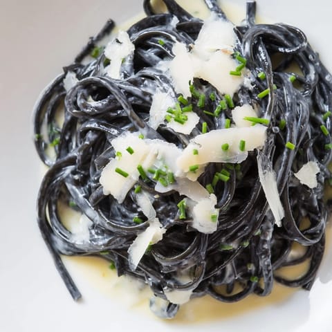 A plated serving of Squid Ink Pasta Dramatic topped with grated Parmigiano-Reggiano, lemon wedges, and chives beside a glass of crisp white wine.